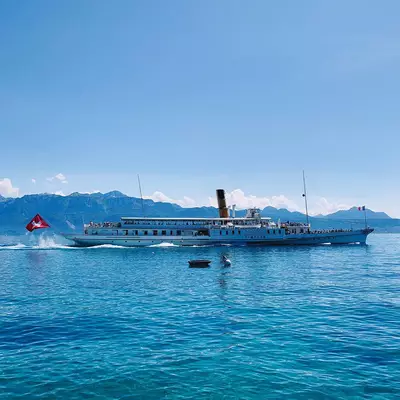 A vintage paddle steamer with a black smokestack and multi-deck design travels through turquoise alpine lake waters, with snow-capped mountains visible along the shoreline under clear blue skies.