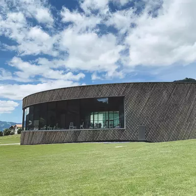 A modern architectural building with a curved dark exterior and geometric pattern sits on a grassy meadow under a blue sky with white clouds, featuring large windows and a minimalist design set against a mountainous backdrop.