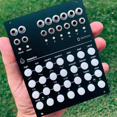 A black synthesizer module held in someone's hand displays two rows of silver jacks at the top, a row of labeled knobs and buttons in the middle including clock, reset, load, and chain controls, and a grid of white knobs below organized by function such as maestro, random, save, square, high, low, mute, and various musical notations and time signatures.