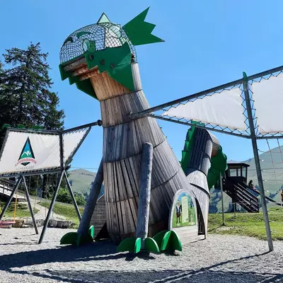 A whimsical green and brown rocket-shaped playground structure with a star on top stands in a grassy area surrounded by safety netting and a trampoline. The structure features circular portholes and climbing elements designed for children's play. Pine trees and clear blue sky frame the outdoor recreational installation.