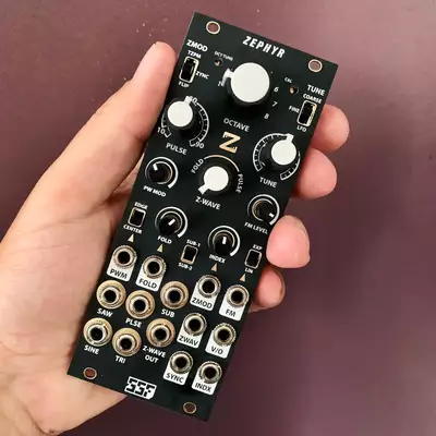 A hand holds a black Eurorack synthesizer module labeled Zephyr with multiple white knobs, switches, and gold patch jacks arranged vertically. The module contains controls for various synthesis parameters including octave, waveform, and tuning functions across its compact faceplate.