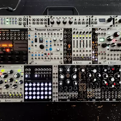 A complex modular synthesizer setup with multiple Eurorack modules arranged across a black case, featuring various knobs, sliders, patch cables, and illuminated displays in different colors including red, blue, and white. The modules include sequencers, samplers, filters, and utility modules with labels visible including 