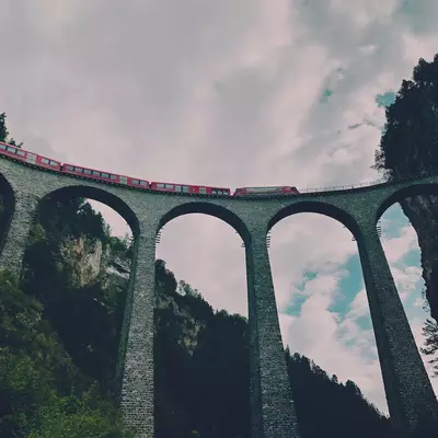 A historic stone viaduct with multiple large arches spans across a forested valley, with a red train traveling on the tracks above the structure against a cloudy sky.