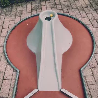 A white plastic violin-shaped object mounted on a terracotta-colored base, photographed from above on a brick patterned surface. The design includes curved sides, a central ridge, and what appears to be mounting hardware at the top.