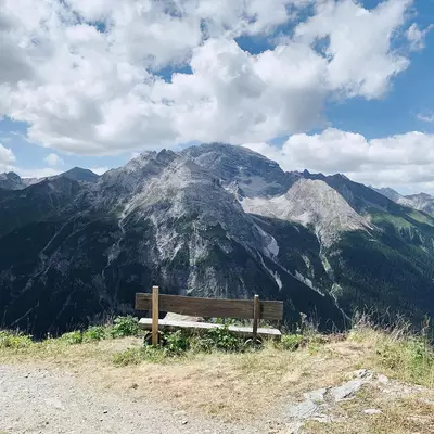 A wooden bench sits on a gravel path overlooking a dramatic rocky mountain peak with forested slopes. White clouds drift across a blue sky above the Alpine landscape. The bench provides a viewing spot facing the imposing gray and dark green mountainside.