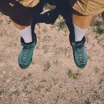 Person wearing white socks and teal suede hiking boots standing on dusty ground with sparse grass, viewed from above.