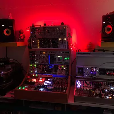A dark music production setup with two studio monitors on stands flanking a central desk, illuminated by red ambient lighting. Multiple synthesizers, drum machines, and control surfaces with illuminated buttons and screens are arranged across the workspace. Audio cables and equipment are visible throughout the setup.