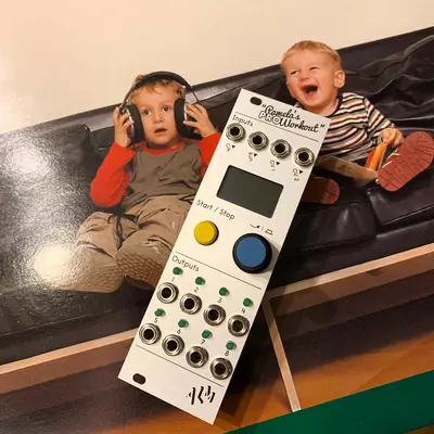 Two young children sit on a black couch holding headphones near a large white electronic music workstation module with a display screen, yellow and blue buttons, and multiple knobs and controls. The child on the right is laughing with an excited expression while the child on the left appears surprised or amazed. The module is propped up on a stand in front of them.
