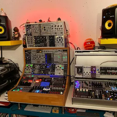 A modular synthesizer setup with two tall Eurorack cases in the center, flanked by yellow-mounted studio monitors on either side. A turntable sits to the left, while various synthesizer modules and controllers are arranged on the desk surface below. Red accent lighting illuminates the wall behind the equipment.