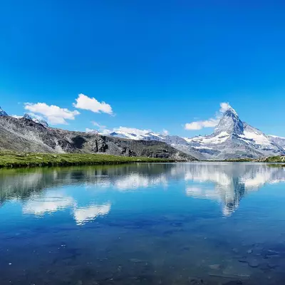 A serene alpine lake mirrors snow-capped mountain peaks and a clear blue sky dotted with white clouds, surrounded by green meadows and forested slopes.