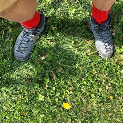A person wearing dark athletic shoes with red socks stands on grass dotted with small yellow flowers, viewed from above showing their feet and lower legs.