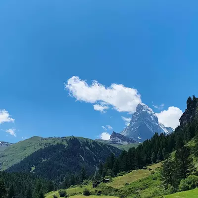 A dramatic mountain peak rises above a lush green valley dotted with evergreen trees and a small wooden structure, under a bright blue sky with wispy white clouds. The steep rocky summit contrasts with the verdant alpine meadows in the foreground. This Alpine landscape appears to be taken during summer in a mountainous region.