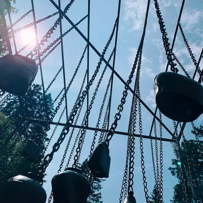 Swings with heavy chains hang against a bright sky with sun glare, surrounded by green foliage, photographed from below looking upward.