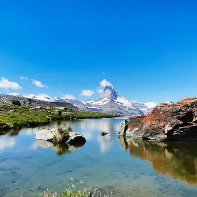 A pristine alpine lake reflects snow-capped mountains and a distinctive peaked summit under clear blue sky, with rocky outcrops and green meadows along the shoreline.
