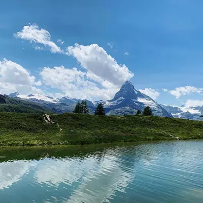 A turquoise alpine lake reflects snow-capped mountains and white clouds under a blue sky, with green meadows and scattered evergreen trees lining the shoreline.