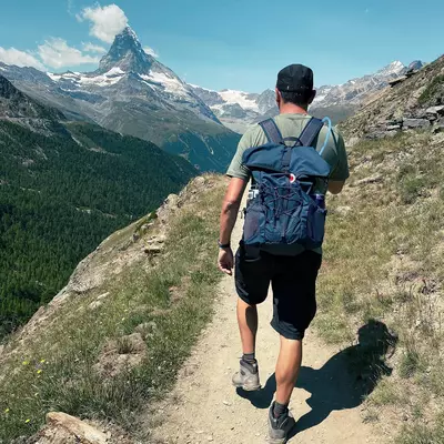 A hiker in a blue backpack and dark shorts runs on a mountain trail with a dramatic peaked mountain in the background, surrounded by forested valleys and alpine terrain.