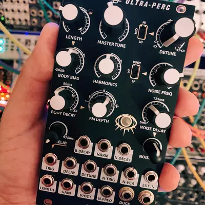 A hand holding a black Erica Synths Ultra-Perc drum synthesizer module displaying twelve knobs for parameters like Length, Master Tune, Decay, Boost Bias, Harmonics, and Noise Freq, with multiple button controls at the bottom, photographed against a blurred background of other synthesizer modules.