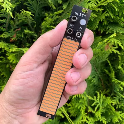 A hand holding a small black Eurorack synthesizer module with an orange wavy pattern stripe, green foliage visible in the background.