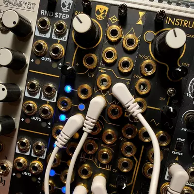 Two white patch cables connected to gold-plated jacks on a dark synthesizer module with multiple knobs, switches, and illuminated blue indicators, part of a larger modular synth setup.