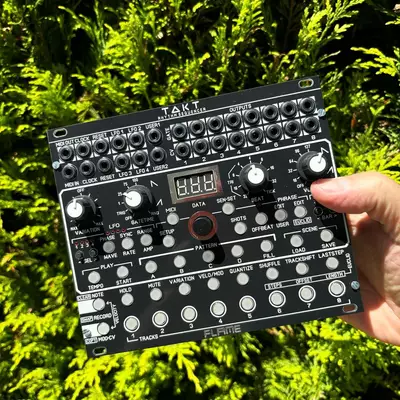 A compact black electronic sequencer/looper device with multiple rows of small buttons, knobs, a digital display, and input/output jacks is held in someone's hand against a bright green foliage background.