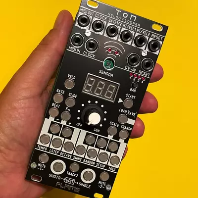 A hand holds a black Eurorack synthesizer module against a bright yellow background, displaying multiple knobs, buttons, and a digital display screen for sound synthesis and modulation control.