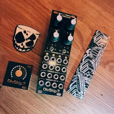 Four DivSkip module packaging items arranged on a wooden surface: a small square box with geometric cat design, a larger rectangular module with multiple knobs and jacks, a small circular sticker, and a patterned rectangular sleeve with chevron designs.
