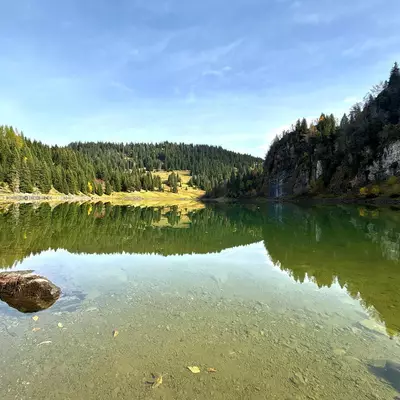 A serene alpine lake with crystal-clear green water reflects surrounding forested mountains and rocky cliffs under a blue sky. Pine trees line the shoreline, with a small clearing visible on the left bank, while a large boulder sits in the shallow foreground water.
