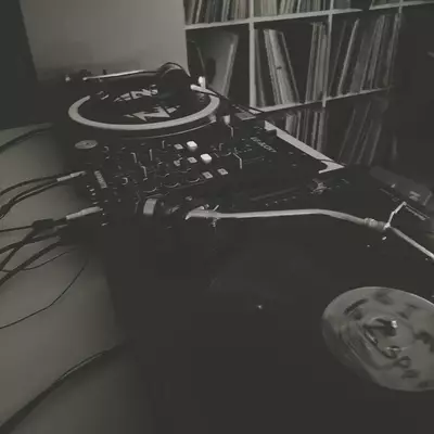 A black and white photograph of a DJ setup featuring a turntable and mixer on a desk, with vinyl records stored on shelves in the background. Cables and equipment are visible in the workspace. The scene captures a music production or DJ station ready for use.