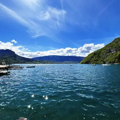 A picturesque view of Lake Annecy, surrounded by lush green mountains under a blue sky dotted with clouds. Boats float on the shimmering water near the shore.