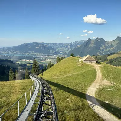 A scenic view of the railway tracks of the Plan Francey funicular, leading to a typical alpine farm building of the Gruyère region, set against a backdrop of distant mountains and a blue sky with scattered clouds.