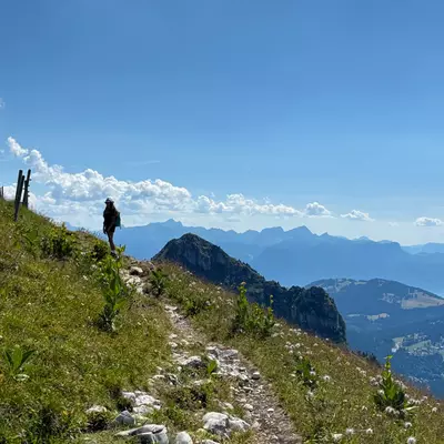 A hiker walking along a mountain path, surrounded by green grass and wildflowers, with distant mountain ranges visible under a bright blue sky.