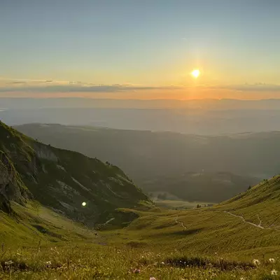 The sun setting over rolling hills, casting golden light across the landscape, with a path trailing between the green slopes.