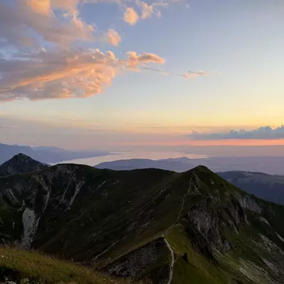 A wide view from the Moléson, showcasing Teysachaux mountain with the sunset illuminating the sky in pink and orange hues. Lake Geneva (Lac Léman) is visible in the distance.