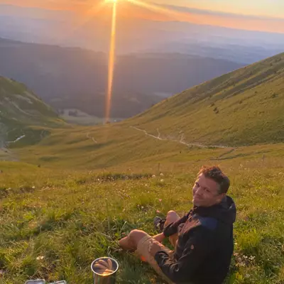 Me having a picnic on a grassy mountainside, with a sunset in the background casting warm light over the landscape. And cooking hot dog sausages