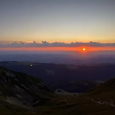 A breathtaking sunset over the Canton of Vaud, with the sun casting a warm orange glow across the horizon, silhouetting the dark mountains below.