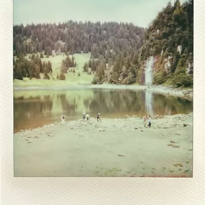 A vintage-style photo of Lac des Chavonnes, depicting a few people at the water's edge with lush greenery and rocky cliffs in the background.