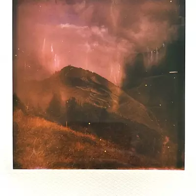 A colored polaroid-style photo of the Saype fresco on the hillside at Grand Chamossaire, under a dramatic sky with hints of sunlight.