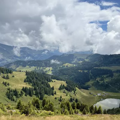 A stunning view from the top of Grand Chamossaire, capturing the rolling hills and lush greenery with Lac de Bretaye visible below, under a sky dotted with dramatic clouds.