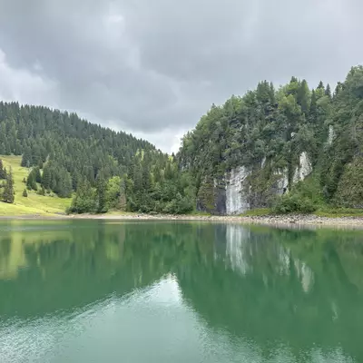 A serene view of Lac des Chavonnes, with calm green waters reflecting the surrounding trees and rocky cliffs under a cloudy sky.