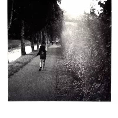 Silhouette of a person running on a tree-lined path along the Broye River, with filtered light creating a play of shadows and contrasts, black and white Polaroid photograph
