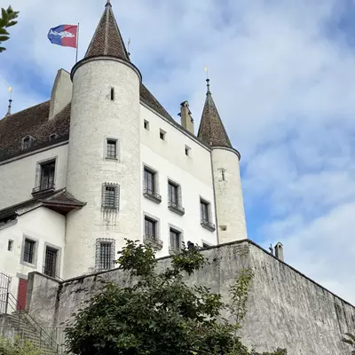 Nyon Castle with two distinctive round towers and conical roofs, Swiss flag flying, against a blue sky with white clouds