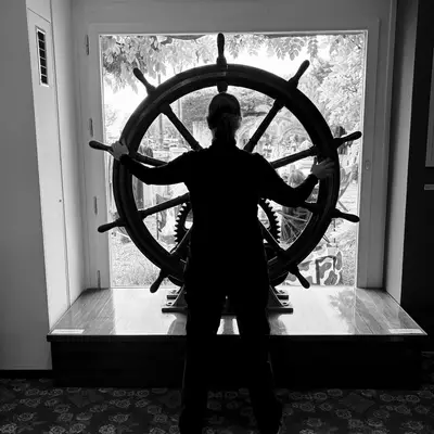 ilhouette of a person standing behind a large ship's wheel at the Lake Geneva Museum in Nyon, with bright windows in the background