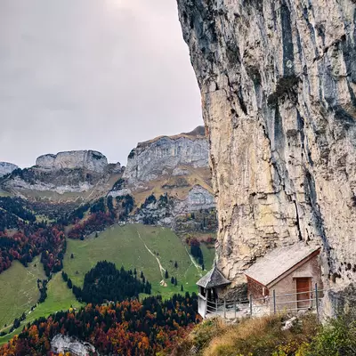 A trail featuring a small wooden walkway clinging to a steep cliff face, with views of green hills and autumn-colored trees.