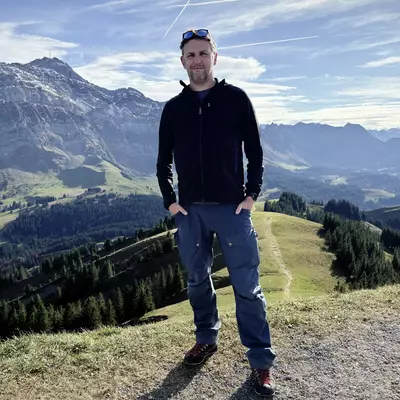Self-portrait at Kronberg summit: man wearing a black hoodie and sunglasses on his head, standing against a dramatic Swiss Alpine panorama featuring layered mountain ranges, green valleys, and forests under a blue sky with wispy clouds