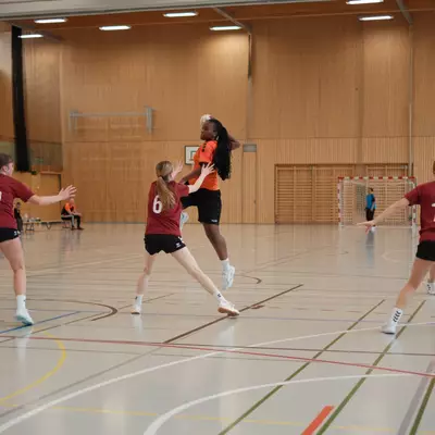 Joueuse du HBC Moudon en maillot orange sautant pour passer le ballon au-dessus des défenseuses du TV Birsfelden numéros 10, 6 et 14 en maillot bordeaux