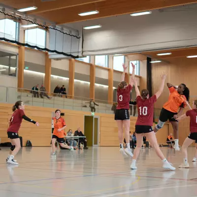 Joueuse du HBC Moudon en maillot orange effectuant un tir en suspension, avec plusieurs défenseuses du TV Birsfelden numéros 3 et 10 tentant de bloquer