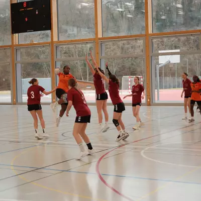 Joueuse numéro 7 du HBC Moudon en orange tirant en suspension, avec le tableau d'affichage indiquant 8-5 et des défenseuses du TV Birsfelden tentant de bloquer