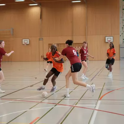 Joueuse du HBC Moudon en maillot orange dribblant le ballon, étroitement marquée par une défenseuse numéro 9 du TV Birsfelden en bordeaux