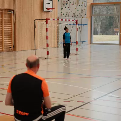 Entraîneur du HBC Moudon portant un gilet orange et noir assis sur un banc, observant la gardienne en bleu debout dans les buts
