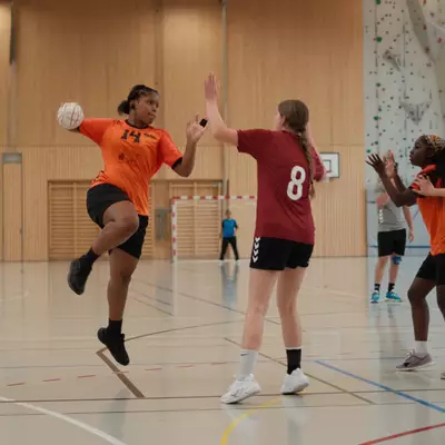 Joueuse numéro 14 du HBC Moudon en orange sautant pour tirer, face aux défenseuses numéro 8 du TV Birsfelden en bordeaux